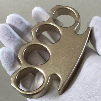 Stonewashed brass knuckle duster held palm-up in a gloved hand, showing four finger holes and clipped front.