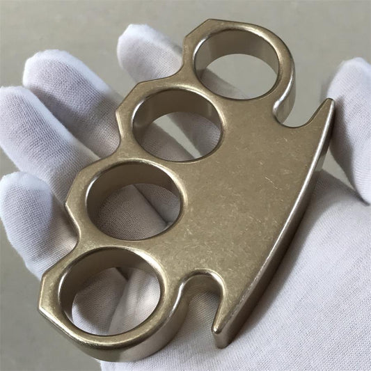 Stonewashed brass knuckle duster held palm-up in a gloved hand, showing four finger holes and clipped front.