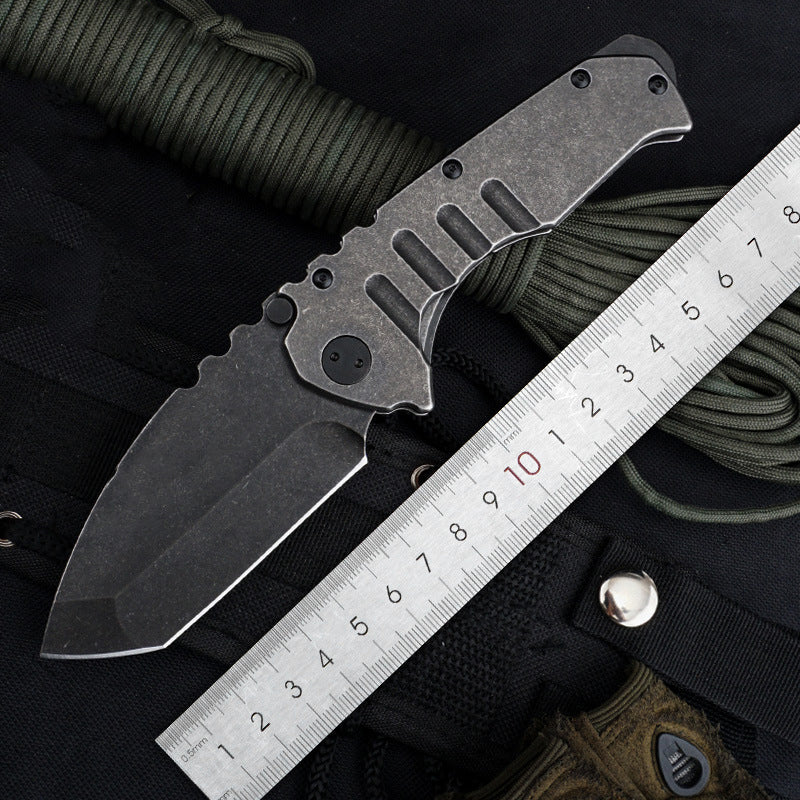 An open folding knife with a dark stonewashed blade and a matching stonewashed steel handle, lying beside a metal ruler on a dark canvas and paracord background, showing elongated lightening slots along the handle.
