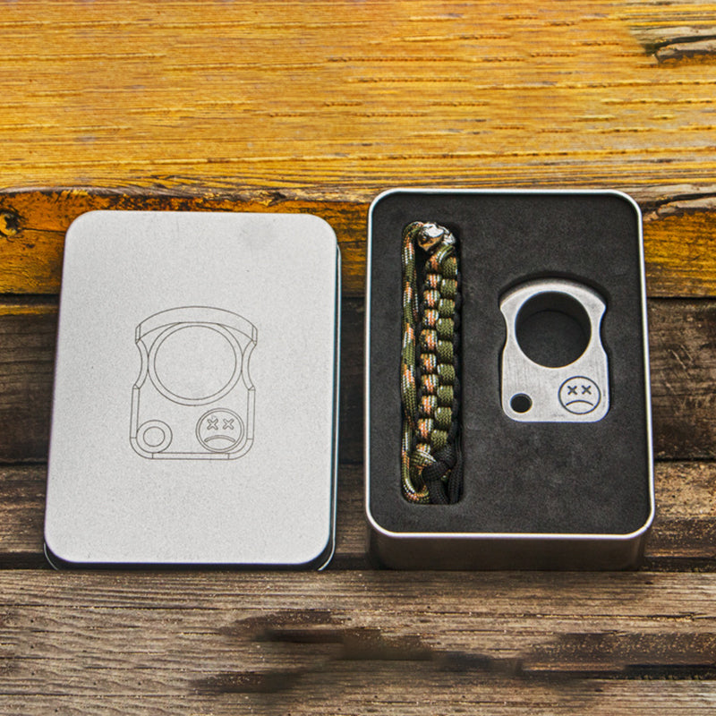 Open metal storage tin with a foam insert holding a dark knuckle weight and a camo braided cord, the lid engraved with the product outline