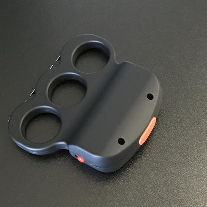 Black three-hole stun device lying on a dark surface with a red side button at the bottom edge and prong points along the left edge