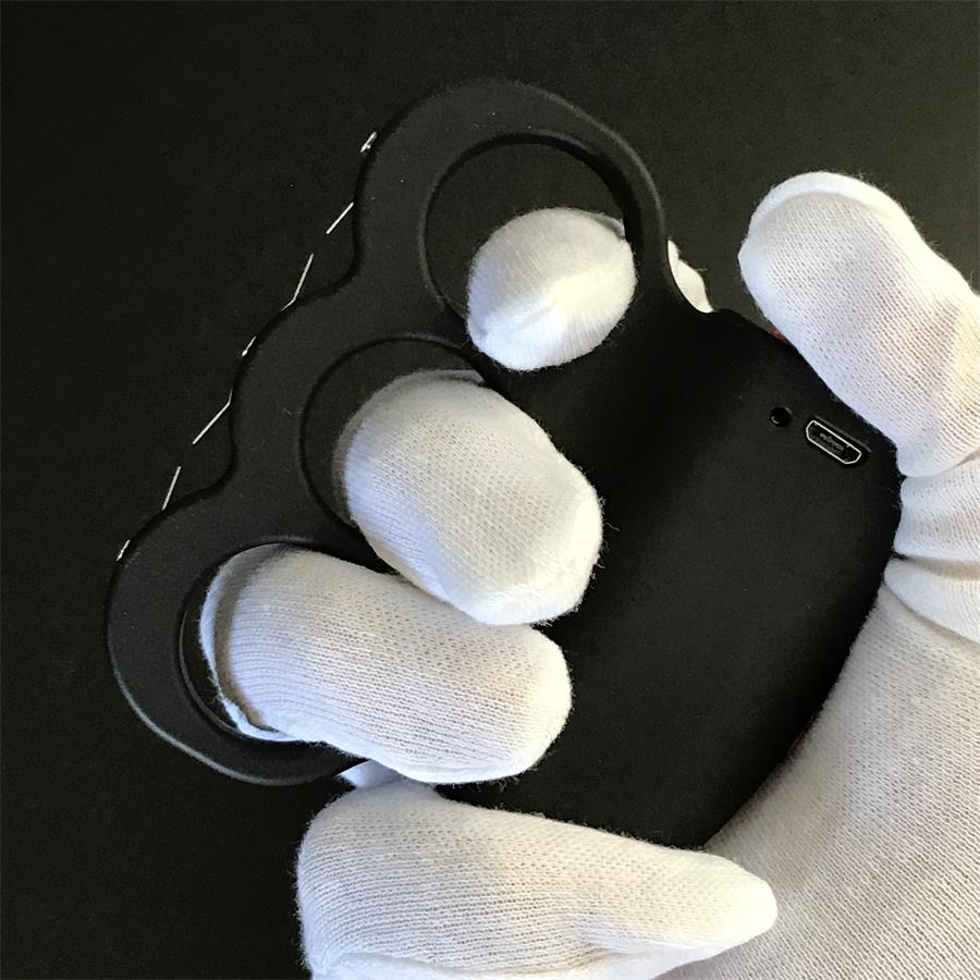 Black three-hole stun device resting on a dark surface, orange-red button on the left side and two small holes on the plate, prong points along the right edge