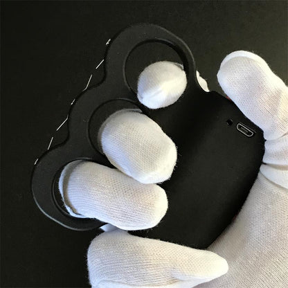 Black three-hole stun device resting on a dark surface, orange-red button on the left side and two small holes on the plate, prong points along the right edge