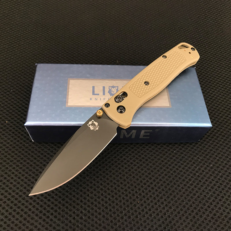Desert tan handled folding knife with black-coated blade and brass pivot accent open on a Liome box