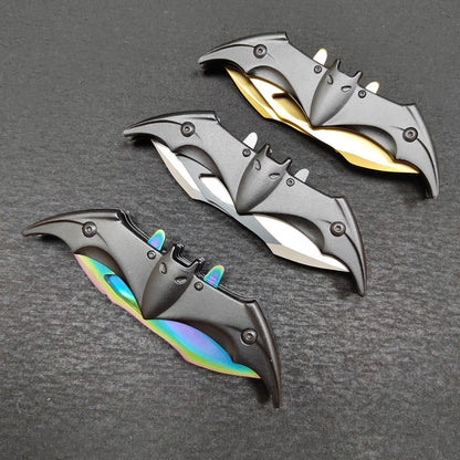 Three bat-shaped folding knives shown closed on a dark gray surface, each with a black bat-head handle cover and exposed wing edges in gold, silver and colourful iridescent finishes, with small cut-outs along the black covers.