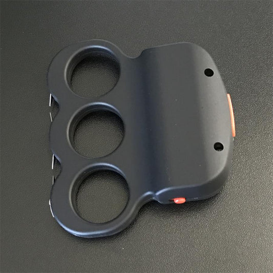 Black three-hole grip stun device lying flat on a dark surface, orange side button and two small holes on the plate, metal prong tips visible along the left edge