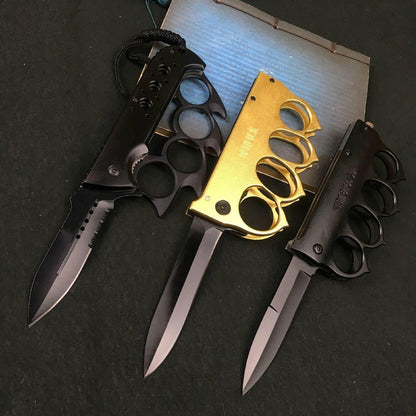 Three knuckle duster folding knives open side by side, gold and black handles over black-coated blades on a dark table