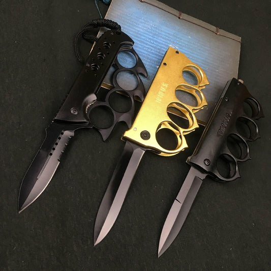 Three knuckle duster folding knives open side by side, gold and black handles over black-coated blades on a dark table