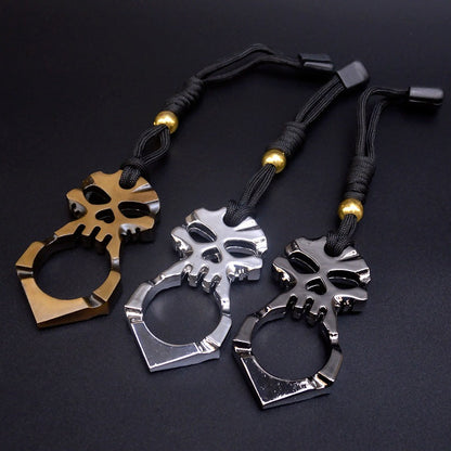 All three finishes laid side by side on a dark surface — gold, silver and titanium black — each skull knuckle duster with its heart-shaped hole and single finger ring, fitted with a black paracord lanyard and gold bead.