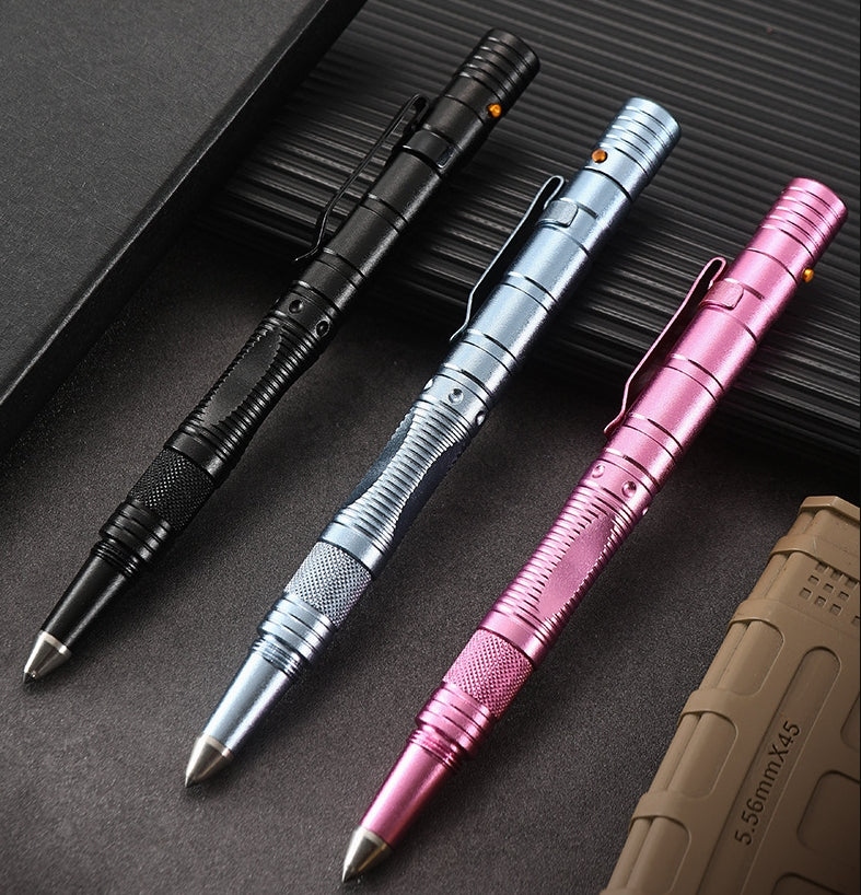 Three tactical pens in black, silver-gray and pink laid diagonally on a dark ribbed surface, shown in side profile with their pocket clips, segmented barrels and knurled grips, beside a tan MOLLE pouch marked 5.56mmX45.