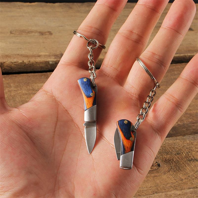 Two mini folding keychain knives rest in an open palm - one open with a silver drop-point blade and one closed - both with navy-and-orange coloured wood handles and a key ring on a short chain.
