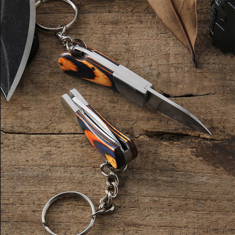 Close-up of two mini folding knives on a wooden table, one open with a slender silver blade and one closed showing its layered spine, both with navy-and-orange coloured wood handles and key ring chains.