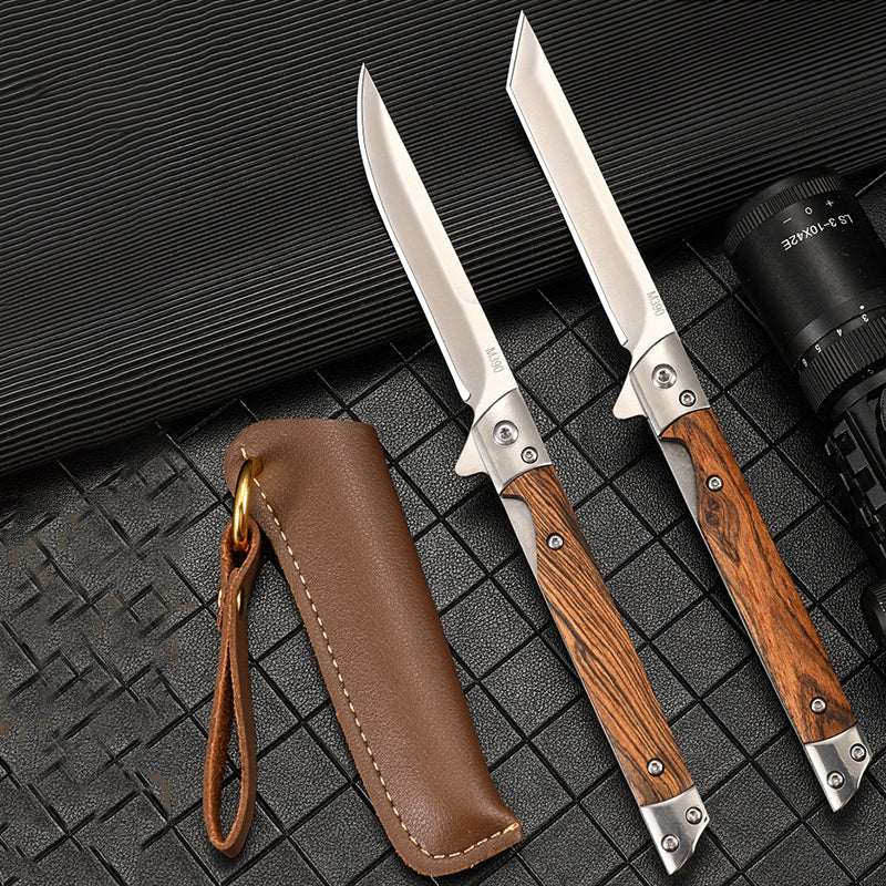 Two open folding knives with bright stainless blades, one drop-point and one angular-tip, above a brown leather sheath with a black flashlight at the right edge