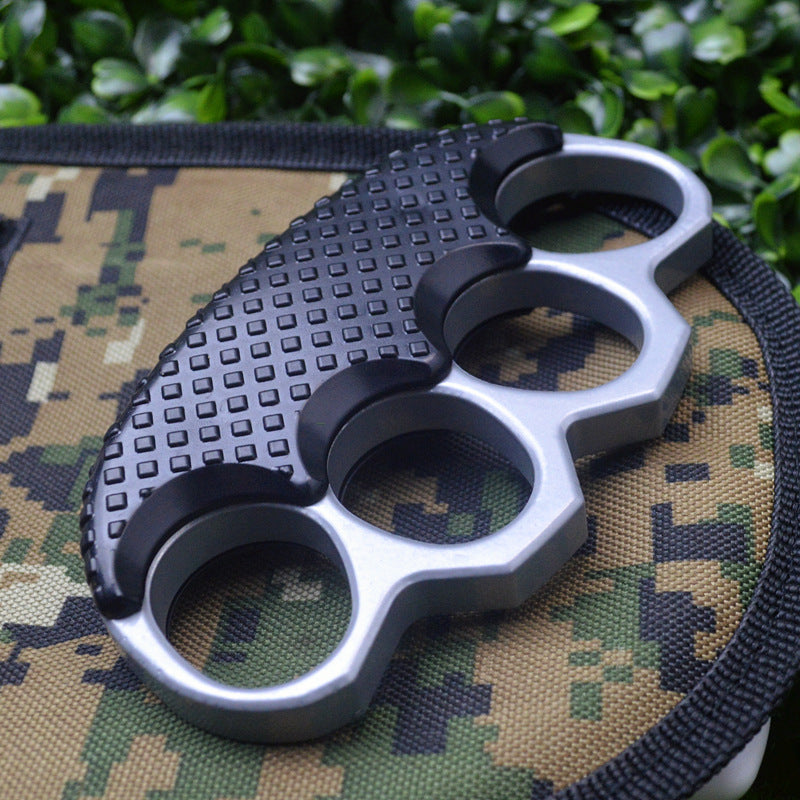 Two-tone knuckle duster with silver gray frame and black studded top panel lying flat on camouflage fabric, four oval finger holes