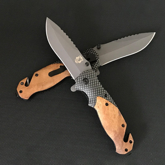 Two Liome X50 folding knives crossed on a dark mat showing wood scales, carbon fiber pattern bodies and grey sawback blades