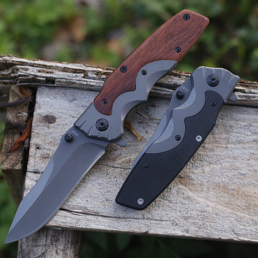 Wood-handle folding knife open with a gray blade and wavy bolster, greenery behind weathered wood