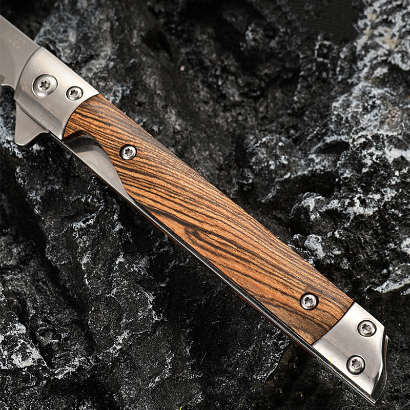 Close-up of an open folding knife showing zebra-striped wood scales, steel pins and bolsters, and the satin blade near the pivot, on dark rock